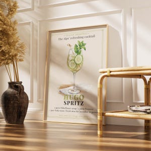 May include: A framed print featuring a watercolor illustration of a Hugo Spritz cocktail. The drink is in a wine glass with cucumber slices and mint. The text on the print reads "The Alps' refreshing cocktail" and "HUGO SPRITZ". The frame is a light brown color.