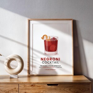 Op de afbeelding: Een ingelijste print met een illustratie van een Negroni-cocktail. Het drankje wordt weergegeven in een glas met ijsblokjes en een garnering. De print bevat de tekst "Negroni Cocktail" en de ingrediënten: gin, vermouth rosso en Campari.