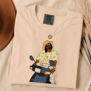 May include: A beige t-shirt with a Comfort Colors label at the neck. The graphic features a person wearing a yellow hat, sunglasses, and a light yellow floral shirt, riding a dark blue scooter. The person wears a white shirt with a pineapple graphic.