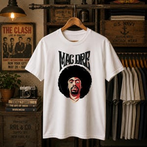 May include: A white short-sleeve t-shirt featuring a graphic print of Mac Dre with a large afro hairstyle. The text "MAC DRE" is printed in a stylized black font above the portrait. The shirt is displayed on a wooden hanger against a dark background.