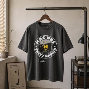 May include: A charcoal gray short-sleeve t-shirt featuring a circular graphic with the text MAC DRE THIZZ NATION surrounding a yellow and black illustration of a person wearing a hat, with the signature Andre printed below the graphic.