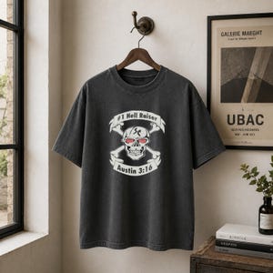 May include: A charcoal gray short-sleeve t-shirt featuring a graphic print of a skull with red eyes and crossed bones. The text #1 Hell Raiser is arched above the skull, and the text Austin 3:16 is printed on a banner below the skull design.