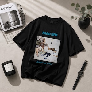 May include: A black short-sleeve t-shirt featuring a graphic print of the Mac Dre California Livin' album cover. The design includes the text MAC DRE in blue block letters above the image and California Livin' in blue script below the image.