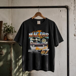 May include: A charcoal grey short-sleeve t-shirt featuring a graphic print of Mac Dre, a vintage car, and flames. The text reads MAC DRE, CALIFORNIA HOT BOY MAKIN' IT SIZZLE!, 67 Cougar, and PLAYBOY, FROM THE BAY BOY. The shirt has a crew neckline.