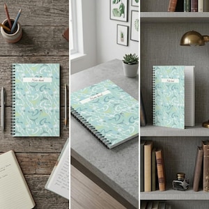 May include: A spiral-bound notebook with a light blue cover featuring a white and green abstract pattern and the text Note book printed on a white label. The notebook has a black wire binding and contains lined paper pages for writing notes.