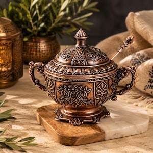 Op de afbeelding: Een koperkleurige metalen suikerpot met bijpassend deksel en een klein lepeltje. De pot is voorzien van ingewikkelde bloemmotieven in reliëf, twee sierlijke handvatten aan de zijkanten en een decoratieve voet. Het deksel heeft een puntige knop.