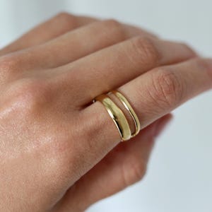 Könnte beinhalten: Zwei goldfarbene Ringe an einem Finger. Ein Ring hat eine breitere, glatte und leicht gewölbte Schiene mit polierter Oberfläche, während der zweite Ring eine dünnere, abgerundete und schlichte Schiene aufweist. Beide haben eine glänzende Oberfläche.