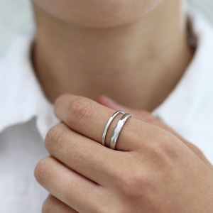 Könnte beinhalten: Ein silberfarbener Ring mit Doppelband-Design. Der Ring besteht aus einem dünnen, abgerundeten Drahtband und einem etwas breiteren Band mit flacher Oberfläche, die parallel zueinander verlaufen und einen geschichteten Look erzeugen.