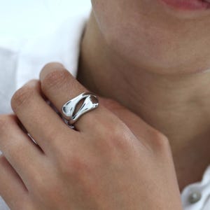 Könnte beinhalten: Ein polierter Ring in Silberoptik mit einer glatten, organischen und fließenden Form sowie einer zentralen Öffnung. Die metallische Oberfläche ist stark reflektierend und bildet eine durchgehende, abgerundete, skulpturale Form.