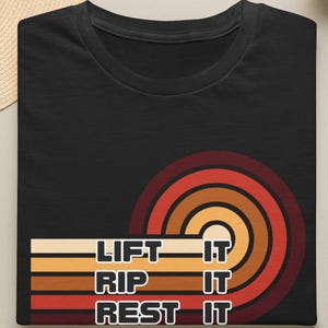 Könnte beinhalten: Ein schwarzes T-Shirt mit einer Grafik aus konzentrischen Halbkreisen in Rot-, Orange- und Gelbtönen. Der Text "LIFT IT", "RIP IT" und "REST IT" ist in weißen Blockbuchstaben über den horizontalen Streifen des Grafikdesigns aufgedruckt.
