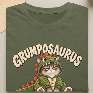 以下が含まれることがあります： オリーブグリーンのTシャツで、赤いトゲのある緑色の恐竜の着ぐるみを着た不機嫌そうな猫のグラフィックが描かれています。猫の上には、クリーム色の丸みを帯びたフォントで「GRUMPOSAURUS」という文字が胸の中央にプリントされています。