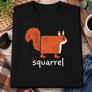 Puede incluir: Una camiseta negra de cuello redondo con un gráfico de una ardilla naranja de forma cuadrada con una cola tupida. Debajo de la ilustración, la palabra "squarrel" está impresa en letras minúsculas blancas sin remates. El diseño está centrado.