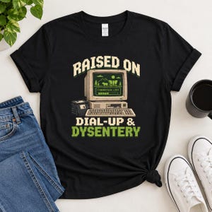 May include: A black short-sleeved t-shirt featuring a graphic of a vintage computer monitor displaying a pixelated landscape and the text "CONNECTION LOST". Below the monitor is a keyboard and the text "RAISED ON DIAL-UP & DYSENTERY".