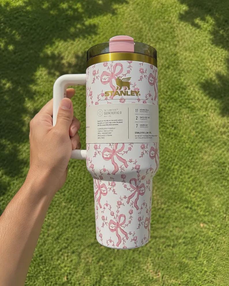 Puede incluir: Un vaso Stanley Flowstate Quencher H2.0 blanco con un patr&oacute;n de lazos y flores rosas, un logotipo dorado, un asa blanca y una tapa rosa. Una etiqueta de producto envuelve el centro, detallando las especificaciones t&eacute;cnicas del art&iacute;culo.