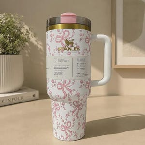 Puede incluir: Un vaso de acero inoxidable blanco con un patr&oacute;n de flores y lazos rosas, con un borde dorado, una tapa rosa y un asa blanca. Una etiqueta de producto envuelve el centro, mostrando el logotipo de Stanley y texto sobre las funciones del Quencher H2.0.