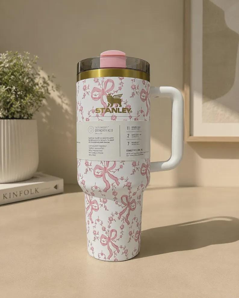 Puede incluir: Un vaso de acero inoxidable blanco con un patr&oacute;n de flores y lazos rosas, con un borde dorado, una tapa rosa y un asa blanca. Una etiqueta de producto envuelve el centro, mostrando el logotipo de Stanley y texto sobre las funciones del Quencher H2.0.