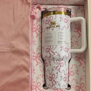 Puede incluir: Un vaso Stanley blanco con un patr&oacute;n de lazos y flores rosas, con borde dorado, tapa rosa y asa blanca. El vaso se presenta en una caja a juego junto a una bolsa de tela rosa con cord&oacute;n que lleva la marca Stanley impresa.