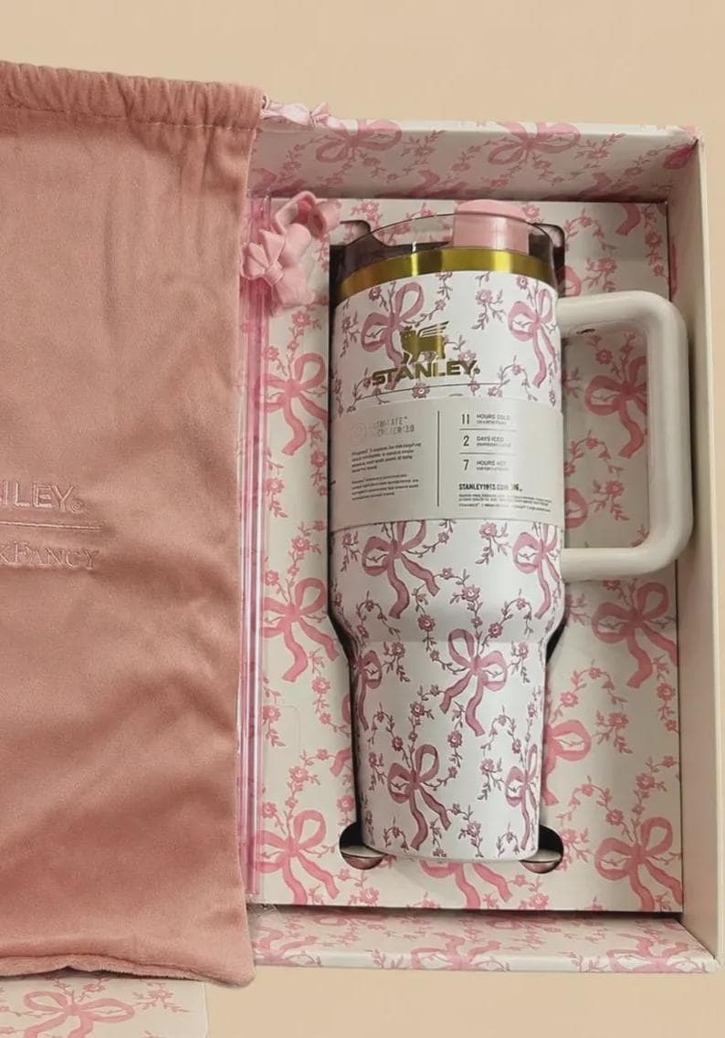Puede incluir: Un vaso Stanley blanco con un patr&oacute;n de lazos y flores rosas, con borde dorado, tapa rosa y asa blanca. El vaso se presenta en una caja a juego junto a una bolsa de tela rosa con cord&oacute;n que lleva la marca Stanley impresa.