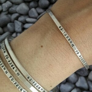 Peut inclure: Trois bracelets en argent grav&eacute;s avec des coordonn&eacute;es. Les bracelets sont empil&eacute;s sur le poignet d'une personne. Les coordonn&eacute;es sont 43.13590 N 70.92640 W.