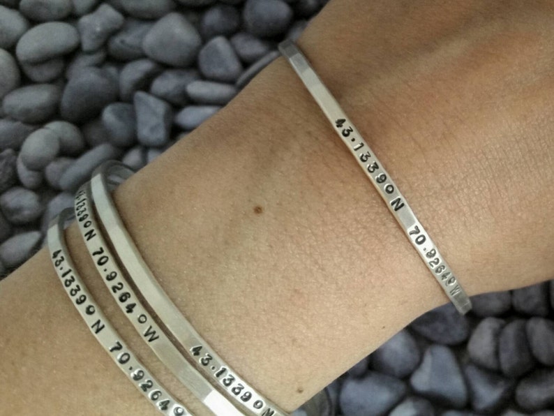 Peut inclure: Trois bracelets en argent grav&eacute;s avec des coordonn&eacute;es. Les bracelets sont empil&eacute;s sur le poignet d'une personne. Les coordonn&eacute;es sont 43.13590 N 70.92640 W.