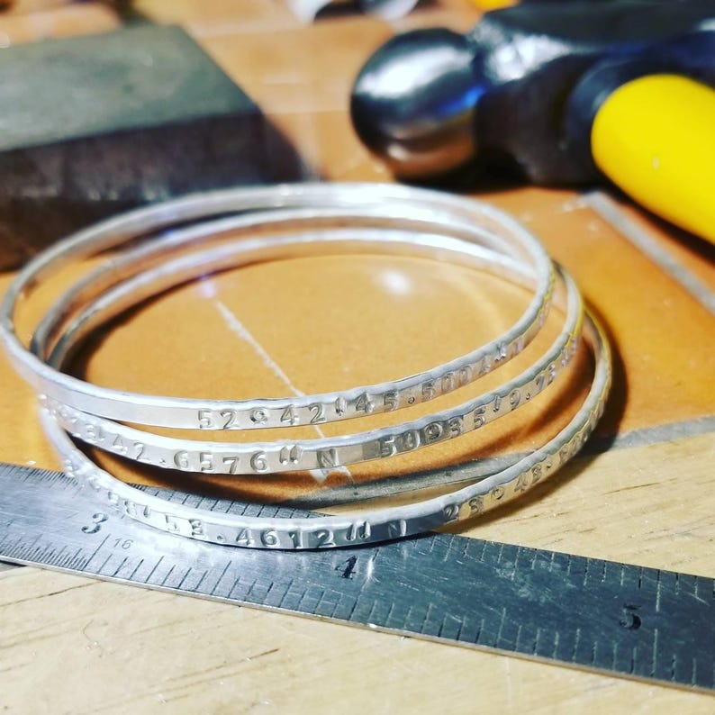 Peut inclure: Quatre bracelets en argent grav&eacute;s de coordonn&eacute;es. Les bracelets sont empil&eacute;s les uns sur les autres et reposent sur une r&egrave;gle en argent.