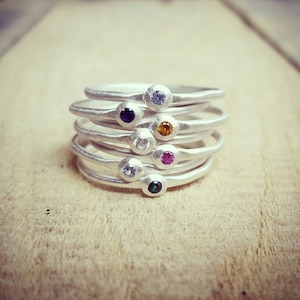 May include: A stack of seven thin silver rings, each featuring a single small round gemstone. The stones are set in a bezel and include colors of light blue, dark blue, yellow, clear, pink, white, and green, arranged in a vertical column.