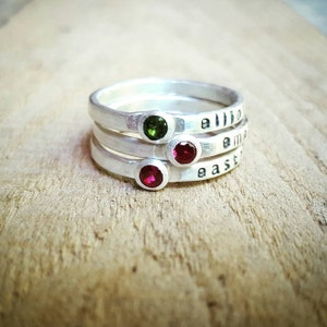 May include: Three silver rings with engraved names "Ellie", "Emma", and "Easton" and small gemstones. The top ring has a green gemstone, the middle ring has a red gemstone, and the bottom ring has a red gemstone.
