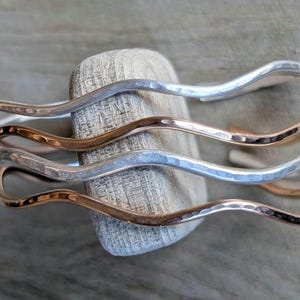May include: Three wavy silver and copper bracelets stacked on top of each other. The bracelets are hammered and have a textured finish.
