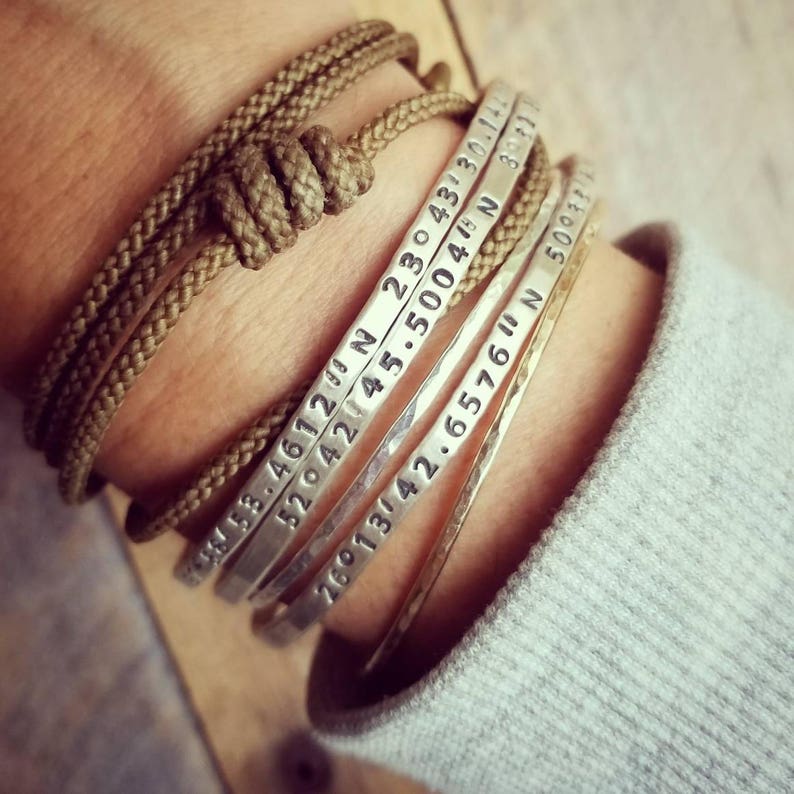 Peut inclure: Un ensemble de quatre bracelets en argent grav&eacute;s avec des coordonn&eacute;es. Les bracelets sont empil&eacute;s sur le poignet d'une personne, avec un bracelet en corde marron sur le dessus.
