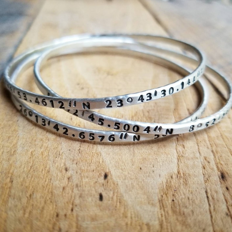 Peut inclure: Trois bracelets argent&eacute;s grav&eacute;s de coordonn&eacute;es. Les coordonn&eacute;es sont 53.4612&deg; N 23&deg; 43' 30.144" N, 26&deg; 13' 42.6576&deg; N, et 45.5004&deg; N.