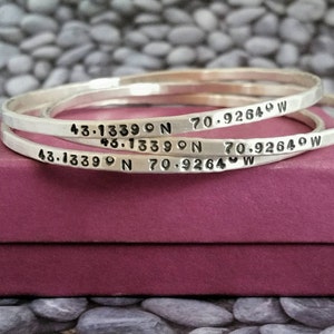 Peut inclure: Trois bracelets argent&eacute;s grav&eacute;s des coordonn&eacute;es "43.13390 N 70.92640 W". Les bracelets sont empil&eacute;s les uns sur les autres et reposent sur une bo&icirc;te rose.