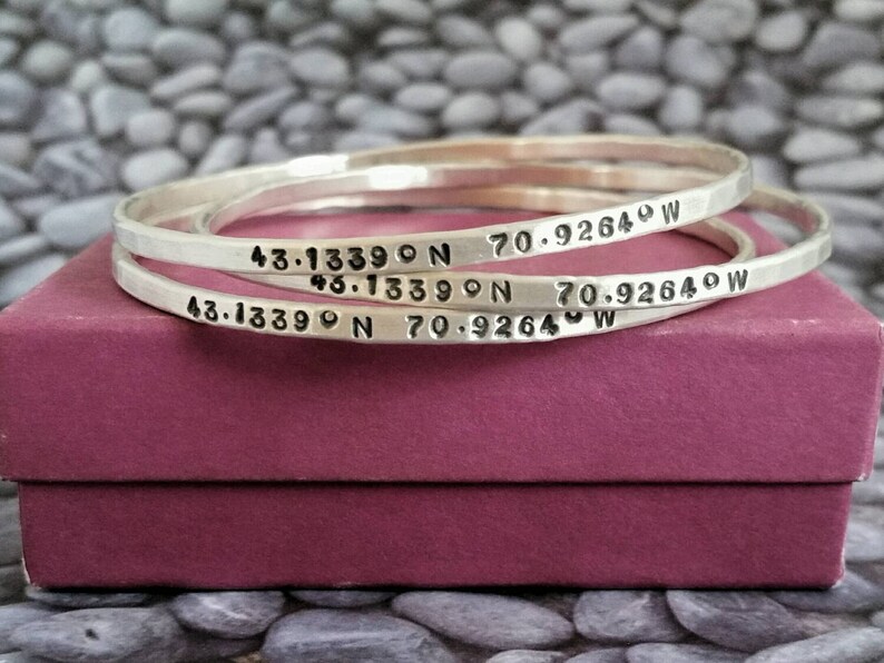Peut inclure: Trois bracelets argent&eacute;s grav&eacute;s des coordonn&eacute;es "43.13390 N 70.92640 W". Les bracelets sont empil&eacute;s les uns sur les autres et reposent sur une bo&icirc;te rose.
