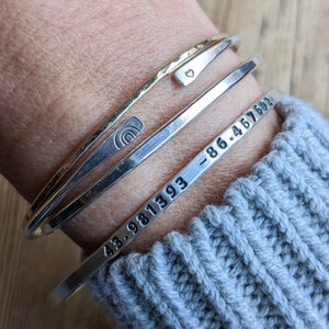 Peut inclure: Trois bracelets en argent empil&eacute;s sur le poignet d'une personne. Le bracelet du haut a un petit charme en forme de c&oelig;ur. Le bracelet du milieu a un petit charme en forme d'arc-en-ciel. Le bracelet du bas a les chiffres "43.981393 -86.467692" grav&eacute;s dessus.