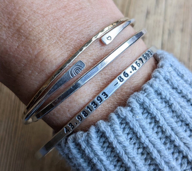 Peut inclure: Trois bracelets en argent empil&eacute;s sur le poignet d'une personne. Le bracelet du haut a un petit charme en forme de c&oelig;ur. Le bracelet du milieu a un petit charme en forme d'arc-en-ciel. Le bracelet du bas a les chiffres "43.981393 -86.467692" grav&eacute;s dessus.