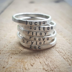 May include: Stack of six silver rings with names engraved on them: Lucia, Sutton, Riley, Rae, Bryler, and Zac.