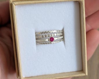 May include: A set of four silver rings displayed in a small, open cardboard box. The rings have different designs, including a twisted rope pattern, a dotted pattern, and a ring with a small, round, red gemstone. The box is lined with white fabric.