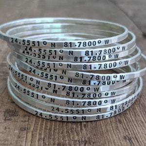May include: A stack of silver bracelets, each engraved with the coordinates "24.5551°N 81.7800°W".