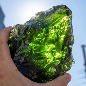 Backlit green obsidian glass crystal glowing bright lime to emerald with sunlight passing through uneven raw edges, showing strong refraction and glossy depth by Evervibes.