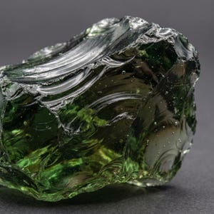 Close view of glossy green crystal glass with curved conchoidal fractures, jagged ridges, and smoky dark edges, highlighting raw green obsidian fracture texture.