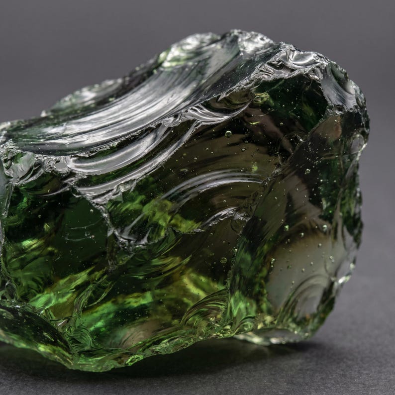 Close view of glossy green crystal glass with curved conchoidal fractures, jagged ridges, and smoky dark edges, highlighting raw green obsidian fracture texture.