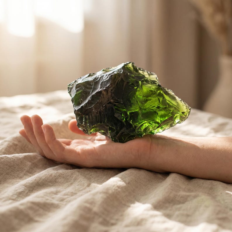Oversized green rough stone held in one hand against daylight, showing its large scale, sharp fractured edges, and emerald transparency for green obsidian rough stone buyers.