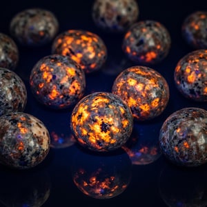 May include: A collection of spherical sunstone gemstones. The stones have a speckled appearance with a mix of gray, brown, and orange hues. The orange areas glow brightly, creating a striking contrast against the darker background.
