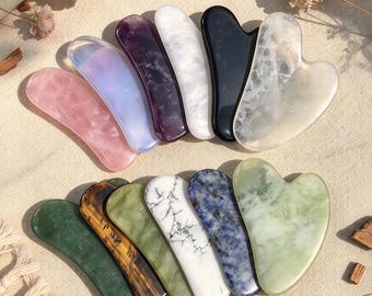 Peut inclure: Une collection de douze outils de massage gua sha en pierre polie et lisse, de formes et de couleurs variées, incluant rose, violet, blanc, noir, vert, marron et bleu, disposés en deux rangées sur une surface en tissu texturé.
