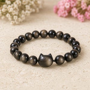 May include: A stretch bracelet composed of round, dark, shimmering obsidian beads and a single central bead shaped like a cat head with pointed ears. The beads feature a metallic gold-toned sheen that catches the light against the dark base.