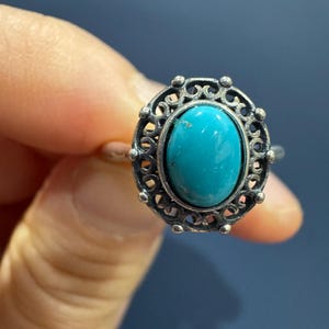 May include: A silver-toned ring featuring an oval-shaped, bright turquoise stone set in the centre. The stone is surrounded by a decorative, openwork metal frame with small, rounded bead accents along the outer edge of the circular setting.