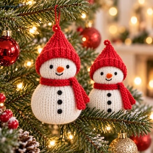 May include: Two white crocheted snowman ornaments with red knitted hats and matching red scarves. Each snowman features a small orange carrot nose, black embroidered eyes, a stitched smile, and three black buttons down the front body.