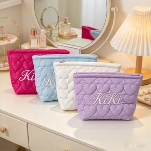 May include: Four quilted cosmetic pouches in hot pink, light blue, white, and lavender. Each pouch features a repeating heart pattern and the embroidered text KiKi in white script on the front. The pouches have a zip closure at the top.