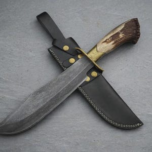 Peut inclure: Un couteau à lame fixe doté d'une lame en acier sombre à motifs, d'une garde en laiton et d'un manche en bois de cerf brun texturé. Le couteau est posé sur un étui en cuir noir avec des coutures blanches et deux rivets en laiton sur la sangle.