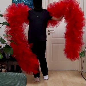 Pode incluir: Uma pessoa segurando dois longos e esvoaçantes lenços de tule vermelho brilhante. Os lenços são franzidos e volumosos, criando um efeito dramático. A pessoa está vestindo uma camiseta preta e calças pretas, com meias brancas.