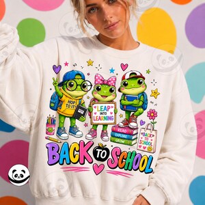 Op de afbeelding: Een crèmekleurig sweatshirt met een afbeelding van drie cartoonkikkers en schoolspullen. De tekst luidt "HOP TO IT!", "LEAP INTO LEARNING", "READ EXPLORE GROW", "BACK TO SCHOOL" en "BACK TO SCHOOL" op een tas met hartjes en sterren.
